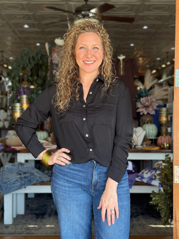 Woman in a black shirt and blue jeans standing in a store with various items in the background.