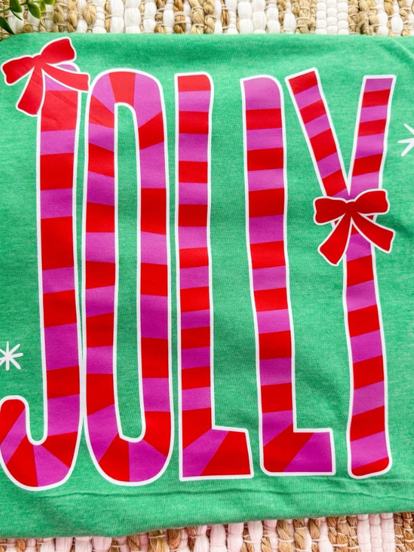 Green pillow with 'JOLLY' in red and pink stripes and a bow on a woven surface.