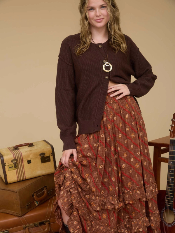 Woman wearing a brown cardigan and patterned skirt standing next to vintage suitcases and a guitar.