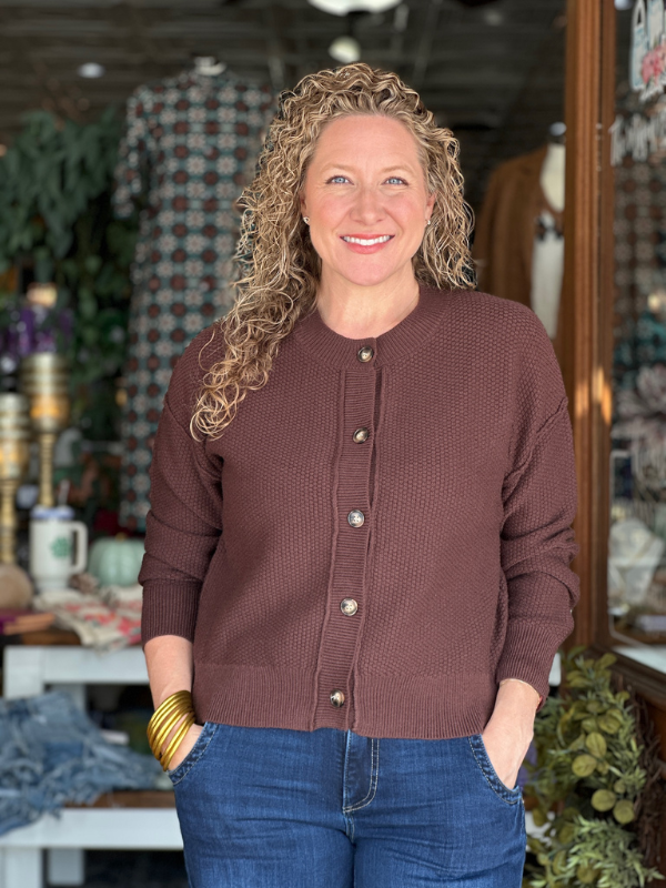 Woman wearing a brown cardigan and blue jeans standing in a store setting.