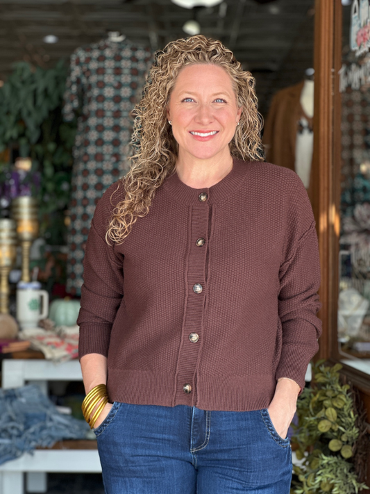Woman wearing a brown cardigan and blue jeans standing in a store setting.
