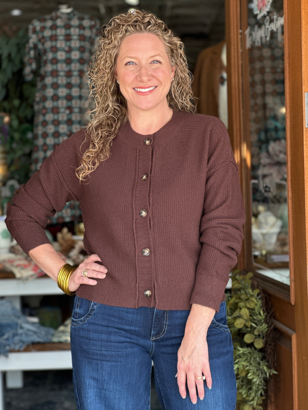 Woman wearing a brown cardigan and blue jeans standing in a store setting.