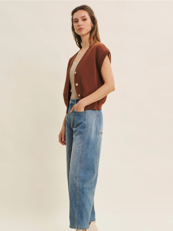 Woman wearing a brown cardigan, beige top, and blue jeans on a plain background