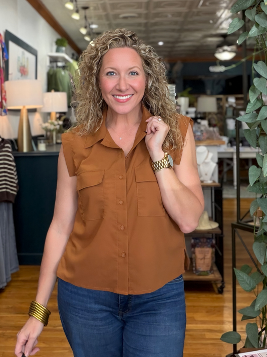 A smiling woman with curly blonde hair stands in a boutique wearing a sleeveless, button-front collared blouse in a warm rust or camel brown color, paired with blue jeans.