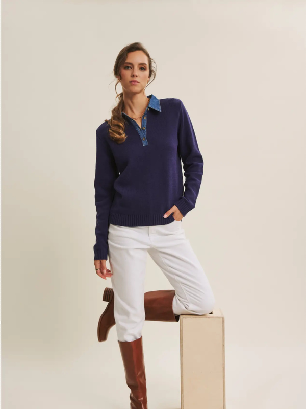 Woman wearing a navy sweater over a blue shirt, white pants, and brown boots on a beige background