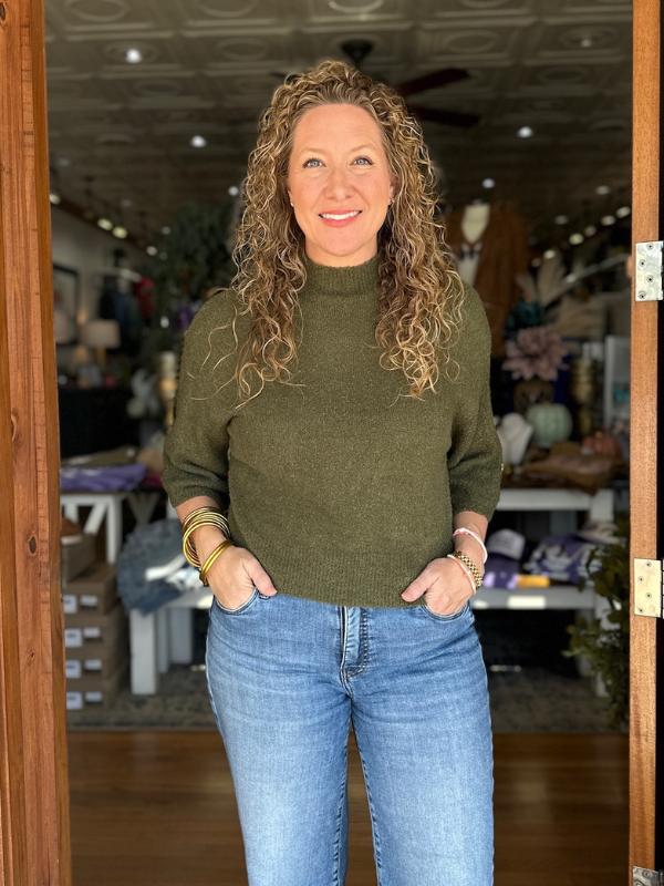 Woman wearing a green sweater and blue jeans standing inside a store.