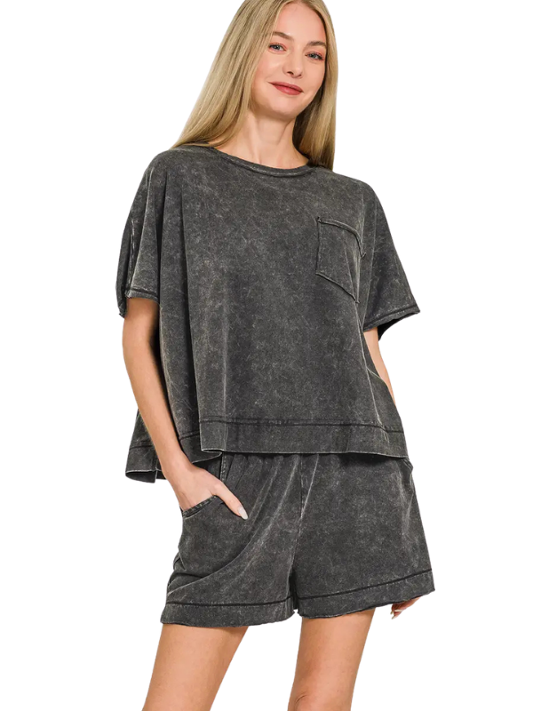 A woman modeling a matching two-piece lounge set in a washed charcoal gray color, featuring a short-sleeve top with a chest pocket and matching shorts, set against a plain white background.