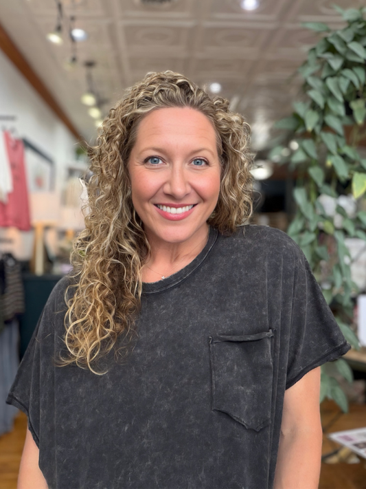 A smiling woman with curly blonde hair wearing a dark gray washed t-shirt with a chest pocket, standing in a retail store with a blurred background.