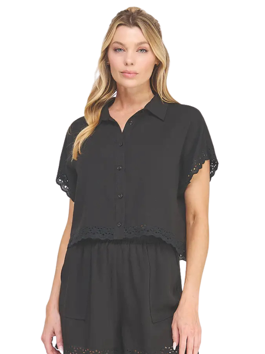 A woman modeling a black short-sleeve button-up blouse with eyelet lace trim on the sleeves and hem, paired with matching black bottoms, against a plain white background.