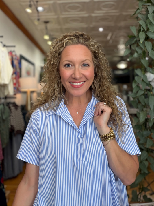 A smiling woman with curly blonde hair wearing a light blue and white vertically striped short-sleeve button-up shirt, standing inside a clothing boutique.