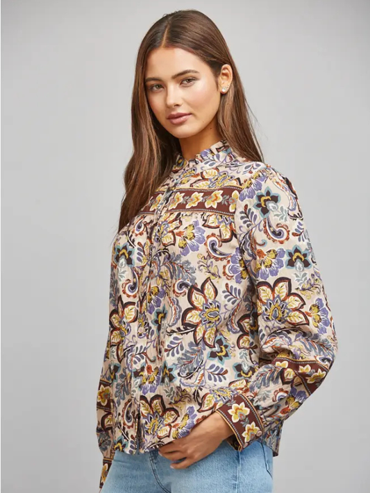 A woman modeling a long-sleeve bohemian-style blouse with a multicolor floral and paisley print on a light beige base, paired with light blue jeans against a solid gray studio background.