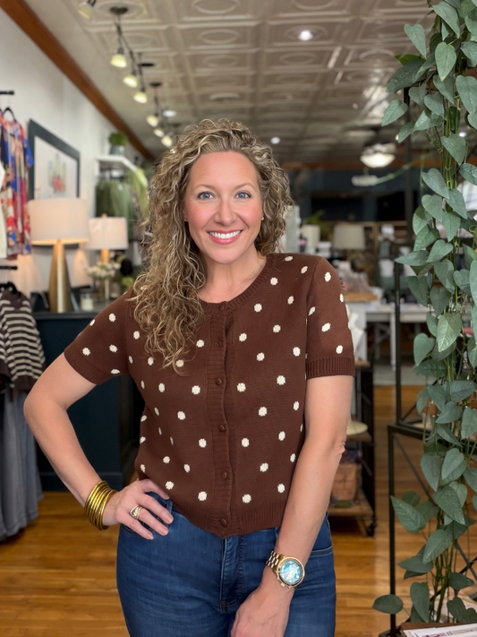 A smiling woman with curly blonde hair stands in a boutique wearing a brown short-sleeve cardigan top with cream polka dots and blue jeans.