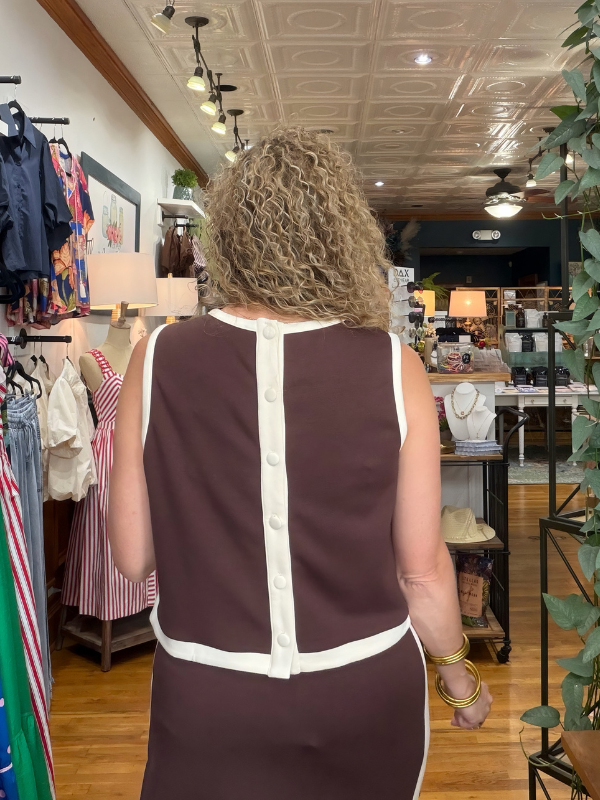 A woman viewed from behind wearing a sleeveless brown dress with white contrast trim and a vertical button placket, standing inside a boutique store.