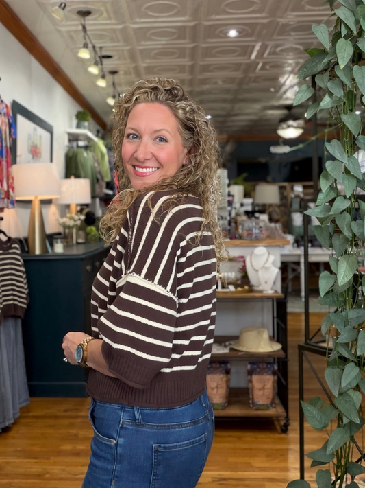 A woman with curly blonde hair stands in a boutique shop, wearing a brown and cream striped knit sweater and blue jeans, looking back over her shoulder.