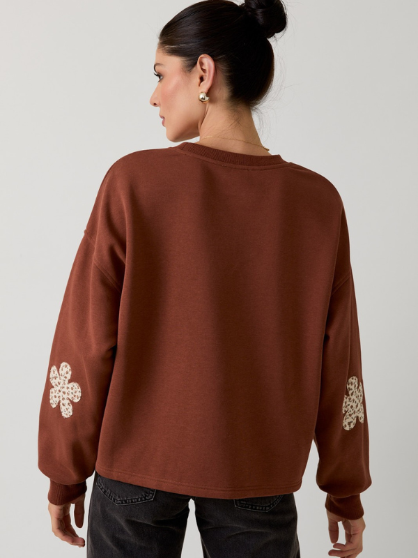 Person wearing a brown sweatshirt with floral designs on the sleeves against a plain background