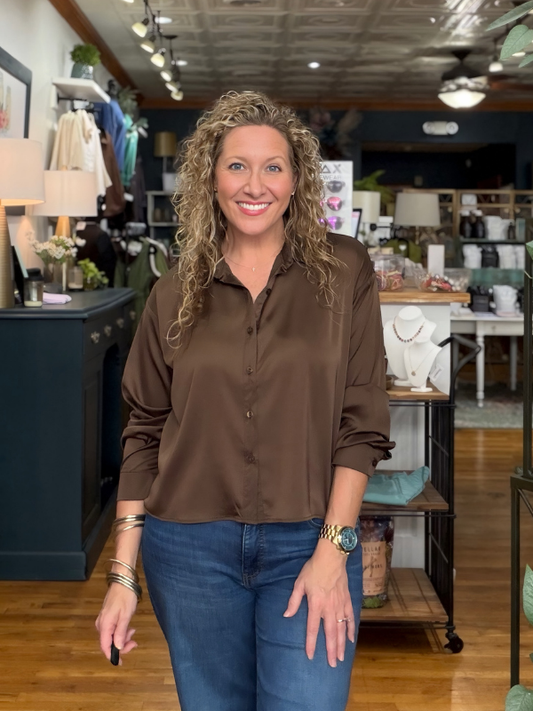 A smiling woman with curly blonde hair stands in a boutique shop wearing a brown button-down shirt and blue jeans.