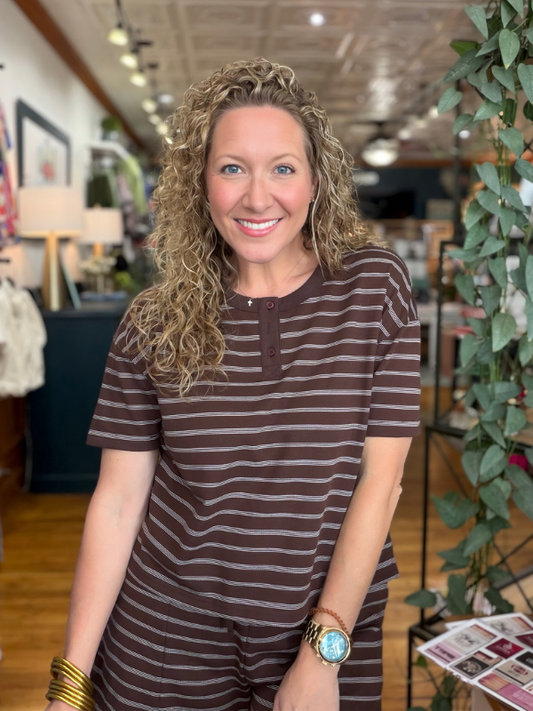A smiling woman with curly blonde hair wearing a brown and gray horizontally striped short-sleeve tunic top, standing in a retail store interior.
