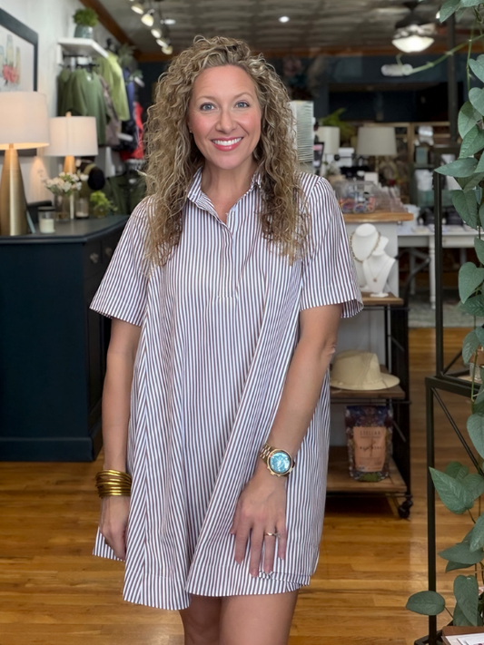 A smiling woman with curly blonde hair stands in a boutique wearing a short-sleeve button-up shirt dress with thin vertical navy and white stripes.