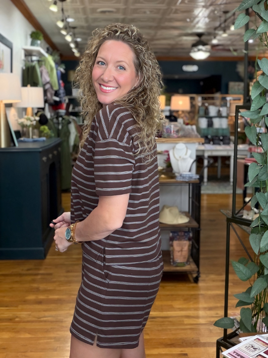A woman with curly blonde hair wearing a brown and beige striped short-sleeve t-shirt dress, posing inside a retail shop.