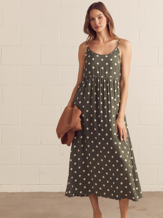 Woman wearing a green polka dot dress holding a brown bag against a white brick wall.