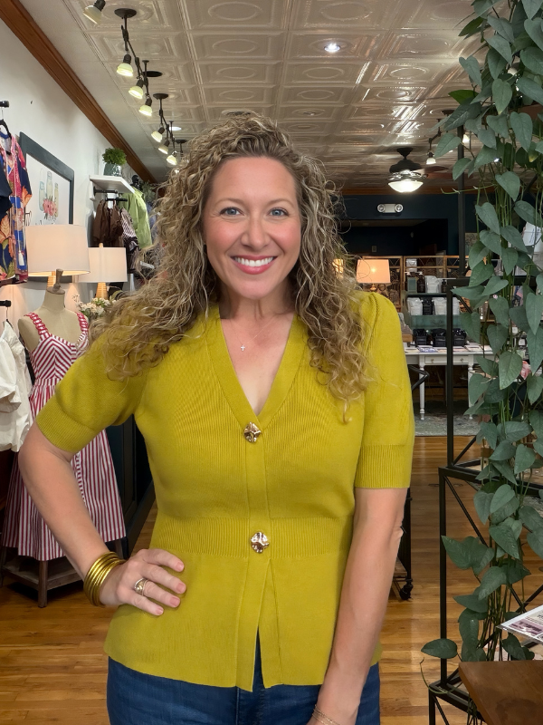 A smiling woman with curly blonde hair stands in a boutique wearing a mustard yellow short-sleeve knit top with decorative gold buttons and blue jeans.