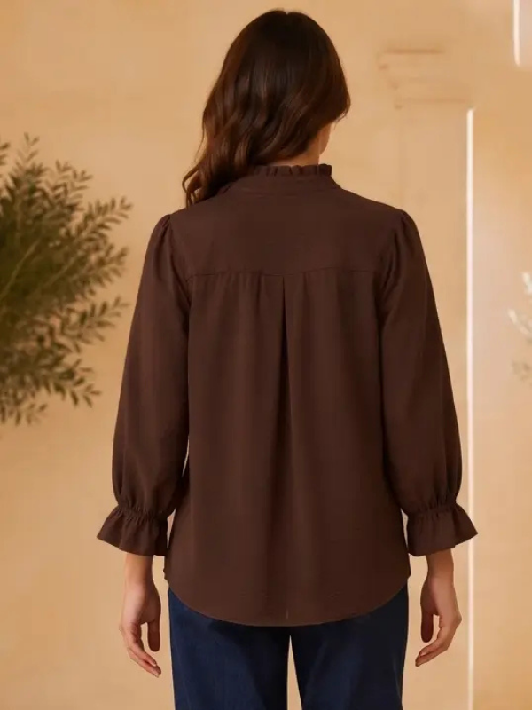 A woman viewed from the back wearing a dark brown long-sleeve blouse with ruffled trim at the collar and cuffs, paired with blue pants, set against a warm beige background with a plant on the left.