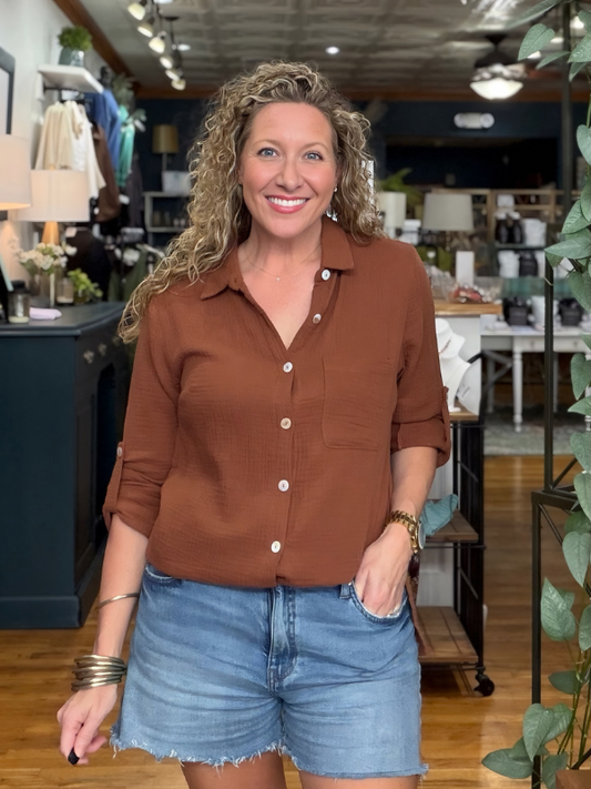 A smiling woman with curly blonde hair stands in a boutique wearing a rust-brown long-sleeve button-up shirt and light-wash denim shorts.