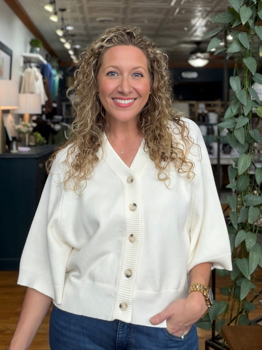 A smiling woman with curly blonde hair wearing a cream-colored knit cardigan with button-front closure and elbow-length sleeves, paired with blue jeans in a boutique setting.