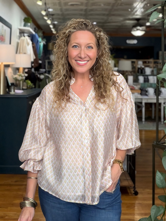 A smiling woman with curly blonde hair wearing a light beige and white patterned blouse with blue jeans, standing inside a retail store.