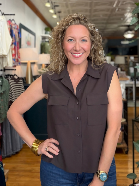 A smiling woman with curly blonde hair stands in a boutique wearing a sleeveless dark brown button-front collared shirt and blue jeans.