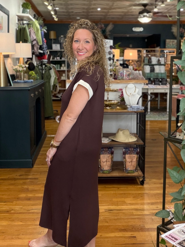 A woman with curly blonde hair stands in a boutique shop, wearing a dark brown sleeveless midi dress with white short sleeves. The shop has wooden floors, clothing racks, and shelves in the background.