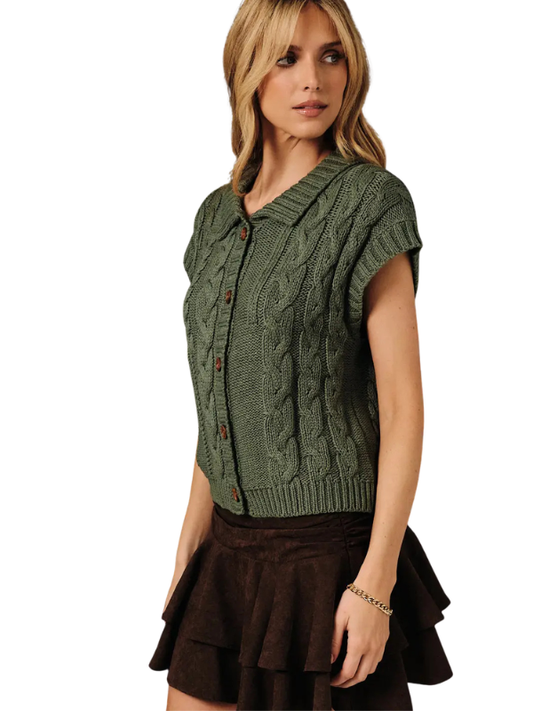 A woman modeling a short-sleeve cable-knit cardigan vest in olive green with brown buttons, paired with a dark brown ruffled skirt, against a plain white background.