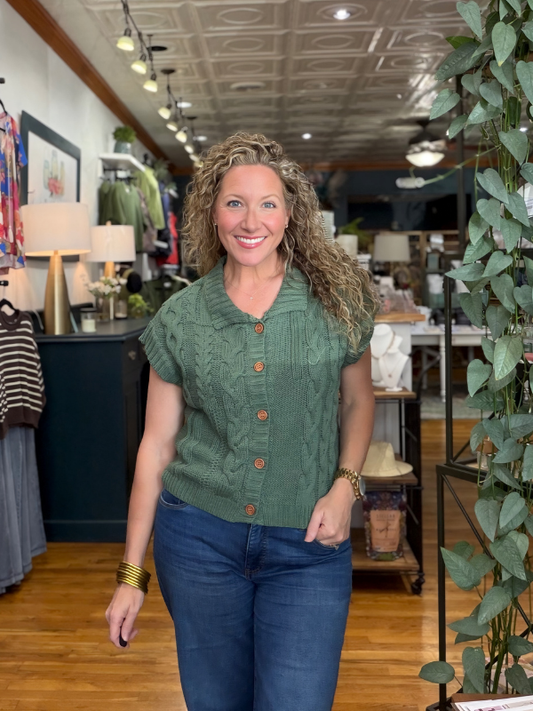 A smiling woman with curly hair stands in a boutique wearing a sage green short-sleeve cable-knit button-front top and blue jeans.