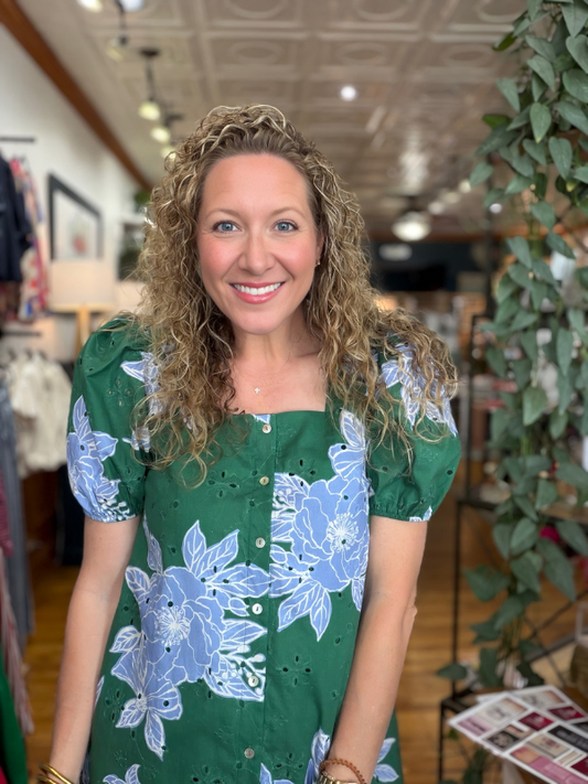 A smiling woman with curly blonde hair wearing a green short-sleeve button-up dress with a light blue floral print, standing inside a boutique.