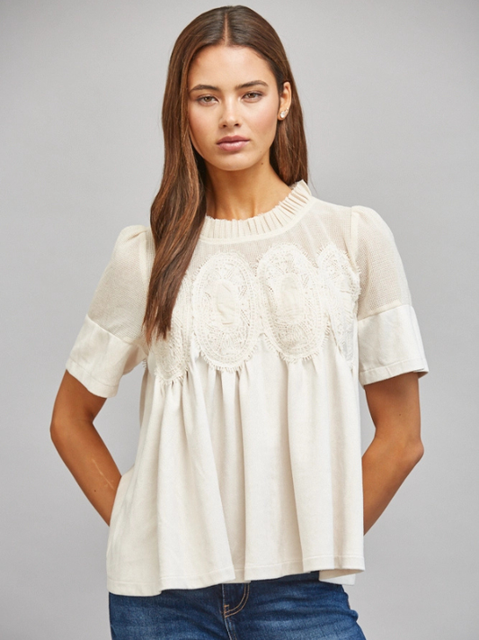 A woman modeling a short-sleeve cream blouse with delicate lace applique across the chest and ruffled detailing at the neckline, paired with blue jeans against a solid gray studio background.