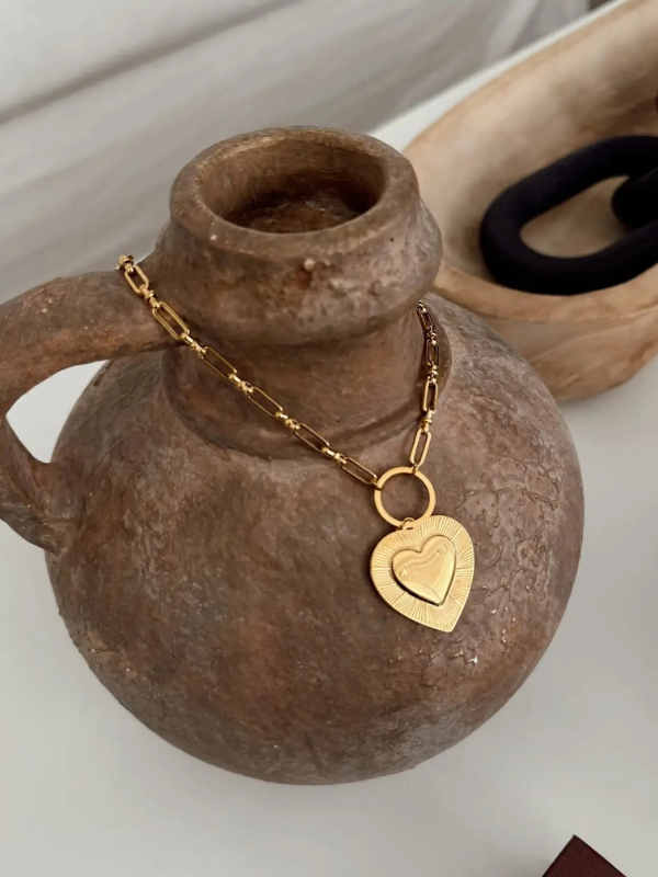 Gold necklace with a heart pendant draped over a brown vase on a white surface.