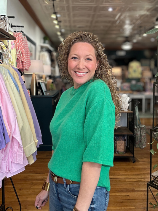 Woman in a green sweater standing in a store with clothing racks and products in the background.