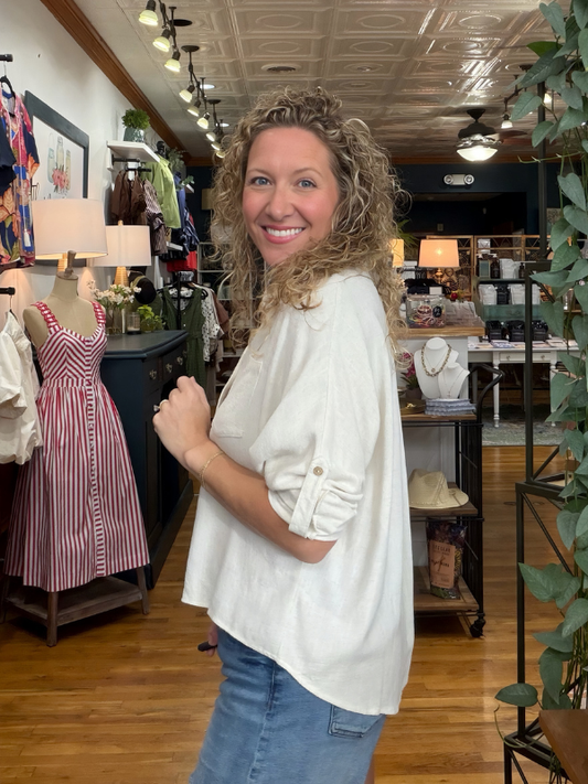 A smiling woman with curly blonde hair wearing an off-white, relaxed-fit blouse and blue jeans stands inside a boutique store. The blouse has a high-low hem and rolled-up sleeves with button tabs.