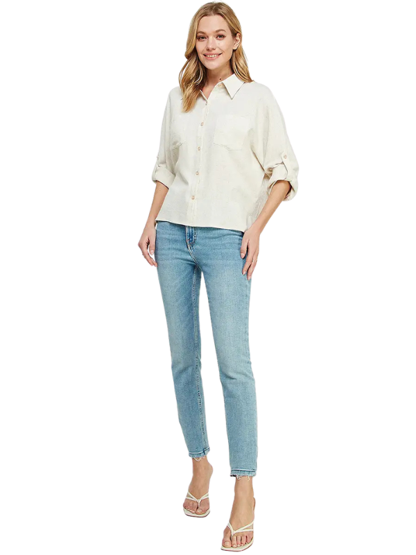 A woman modeling a light cream-colored, long-sleeve button-up shirt with rolled-up sleeves, paired with light-wash blue skinny jeans and clear high-heeled sandals, against a plain white background.