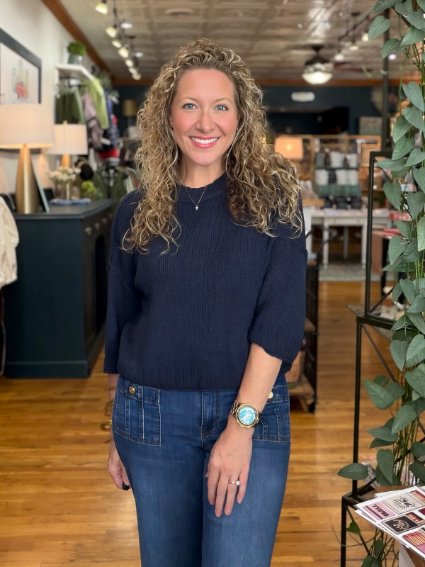 A smiling woman with curly blonde hair stands in a retail store interior wearing a dark navy ribbed knit mock-neck top and blue jeans.