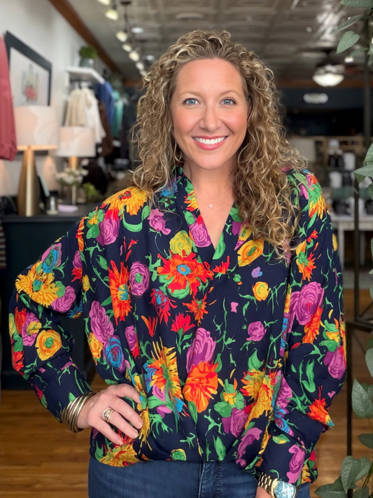 A smiling woman with curly blonde hair wearing a navy blue long-sleeve blouse with a vibrant multicolor floral print, paired with blue jeans, standing in a boutique setting.