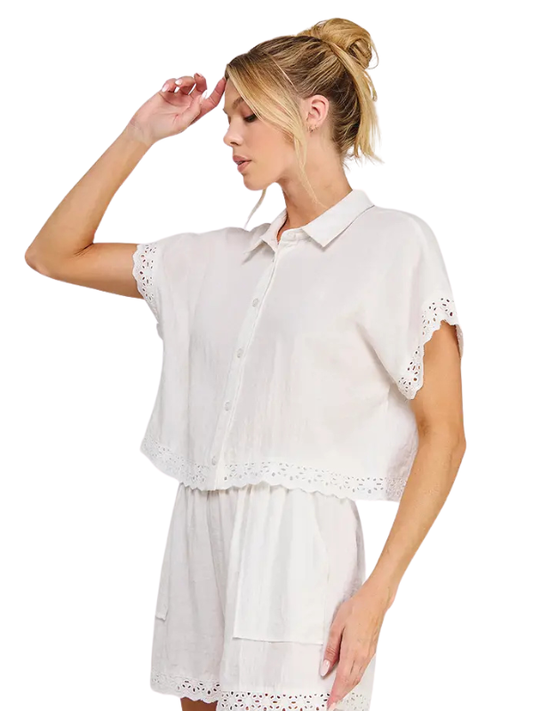 A woman modeling a white short-sleeve button-front shirt with eyelet lace trim, paired with a matching white skirt, against a plain white background.