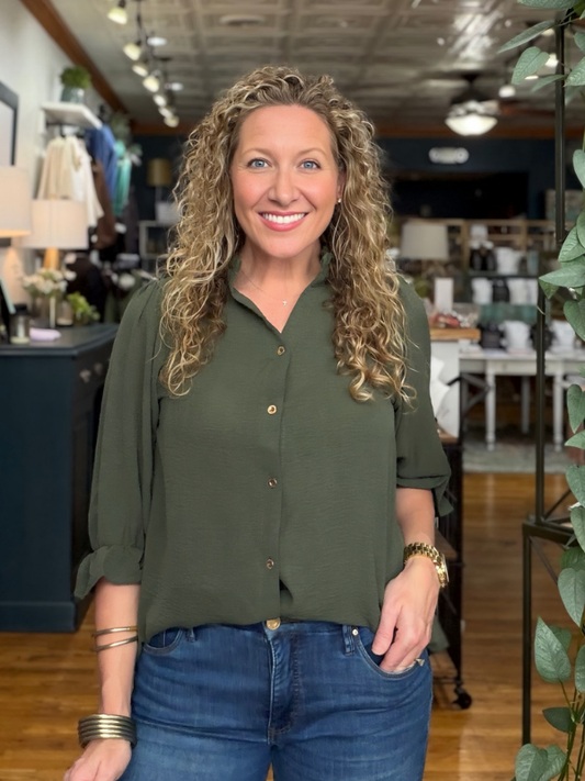 A smiling woman with curly blonde hair wearing an olive green button-down shirt and blue jeans, standing in a retail store environment.