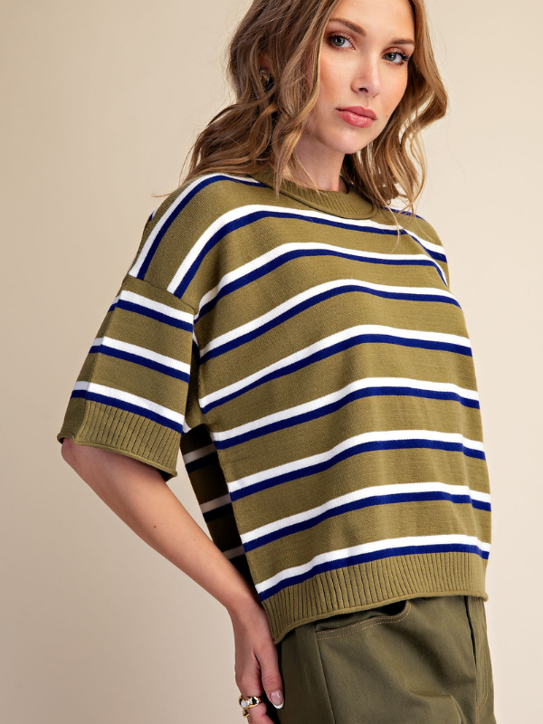 A woman wearing a short-sleeve knit sweater with horizontal stripes in olive green, navy blue, and white, paired with olive green pants, set against a plain beige background.