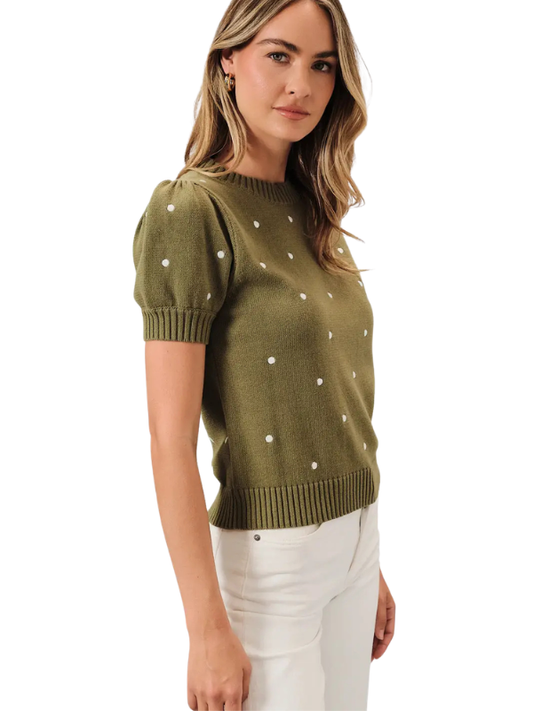 A woman wearing an olive green short-sleeve knit sweater with small white embroidered dots, paired with white pants, shown against a plain white background.