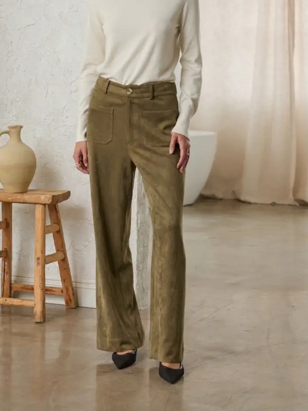 A person wearing olive green wide-leg corduroy pants with a cream long-sleeve top and black pointed shoes, standing in a softly lit indoor room with a stool, vase, and curtain in the background.