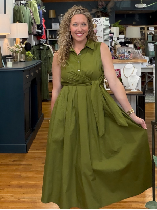 A woman with curly blonde hair wearing a sleeveless olive green midi dress with a button-down collar and gathered waist, standing in a clothing boutique and holding out the side of the dress to show its flow.