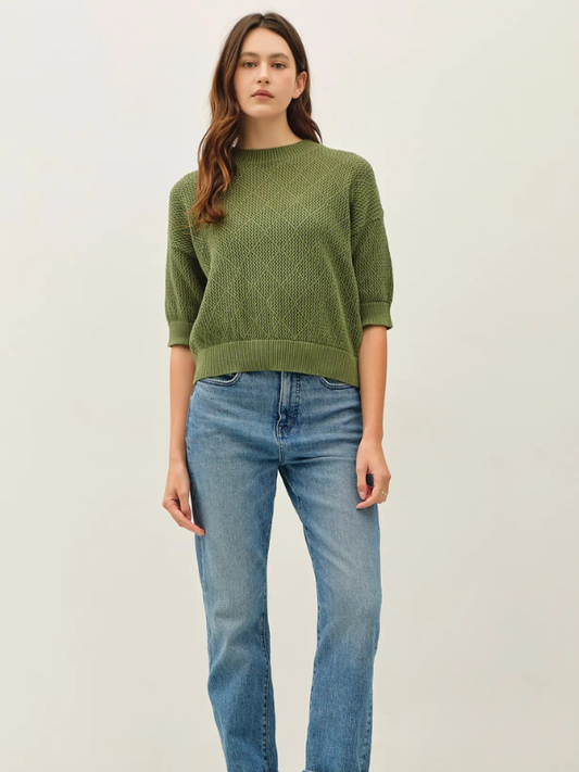 A woman modeling a textured olive green knit sweater with three-quarter sleeves, paired with light-wash blue jeans, against a plain off-white studio background.