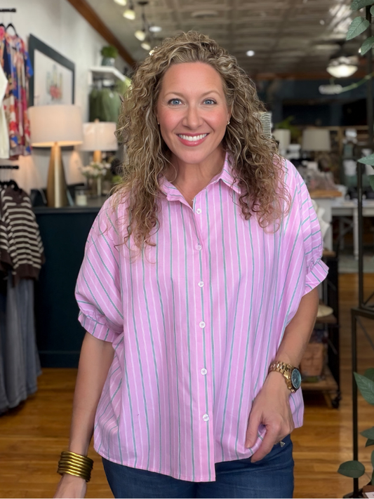 A smiling woman with curly blonde hair wearing a pink and white vertically striped short-sleeve button-up shirt, standing inside a retail store.