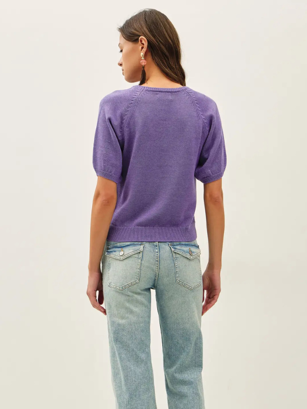 A woman shown from the back wearing a short-sleeve purple knit top and light blue jeans against a plain off-white studio background.